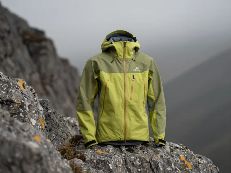 A three-layer hardshell mountaineering jacket on a rocky alpine ridge, dramatic overcast light