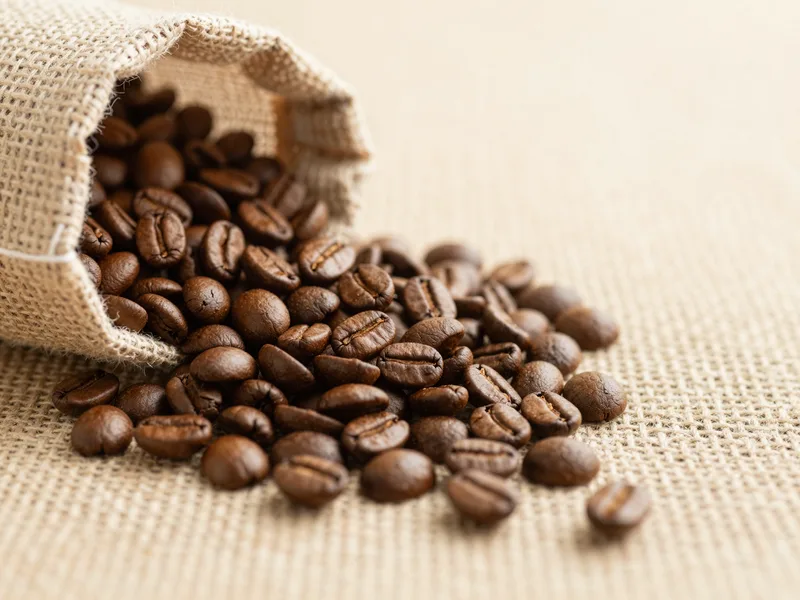 A bag of single-origin coffee beans spilling onto burlap, warm espresso-brown tones, product photo